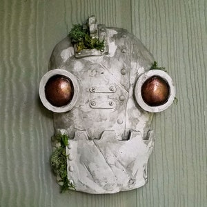 May include: A gray, metal robot mask with two large, round, copper-colored eyes. The mask has a textured surface and is decorated with small, metal rivets. There is green moss growing on the top and sides of the mask.