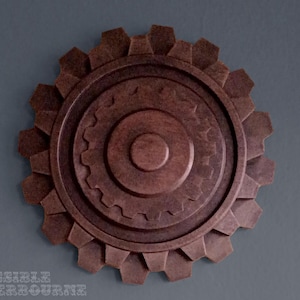 May include: A brown, gear-shaped wall decoration with multiple concentric circular gears. The central gear has a small, raised circle. The outer gear has a series of triangular teeth. The background is a solid gray.