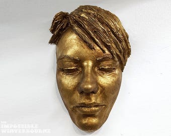 May include: A gold-colored sculpture of a person's face with their eyes closed. The sculpture is made of a textured material that resembles wood.