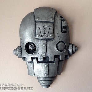 May include: A silver-colored, steampunk-style robot head with visible gears in one eye. The head features riveted plates, a rectangular emblem with the letters "I W", and the text "THE IMPOSSIBLE WINTERBOURNE" at the bottom.