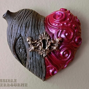 May include: Heart-shaped wall decor, split into two halves. One side is textured gray, the other is swirled red with a metallic sheen. A gold-colored keyhole detail is centered on the heart. The text "THE IMPOSSIBLE WINTERBOURNE" is at the bottom.