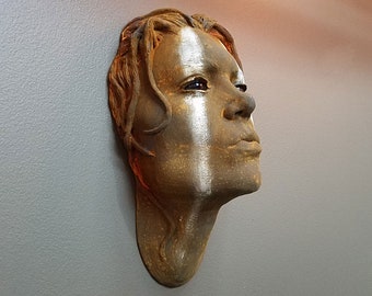 Könnte beinhalten: Eine Keramikskulptur eines menschlichen Gesichts mit weißen Farbakzenten. Die Skulptur ist so gestaltet, dass sie wie verwittertes Metall aussieht.