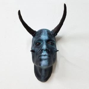 May include: A blue and black sculpture of a face with horns. The face has a serious expression and is painted with a blue and black color scheme.