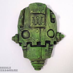 May include: Green, stylized robot head sculpture with rivets and mechanical details. The sculpture has a weathered appearance and features the initials "I W" on the front. The text "THE IMPOSSIBLE WINTERBOURNE" is at the bottom.