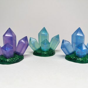 May include: Three clusters of iridescent crystals, each with a different color gradient. The crystals are arranged in a starburst pattern and are set on a green base.