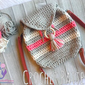 May include: Crochet pattern for a gray, white, and pink striped backpack with a wooden ring and a pink tassel. The backpack is on a wooden surface with yarn balls in the background.