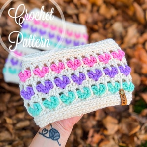Puede incluir: Una diadema de crochet blanca con un patrón de puntadas en forma de corazón en rosa, morado y turquesa. La diadema tiene una etiqueta de cuero marrón que dice "MINI".