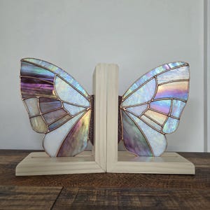 May include: Butterfly-shaped bookends with iridescent stained glass wings in shades of purple, white, and blue. The wings are framed with copper and set on a light wood base. The bookends are designed to hold books upright.