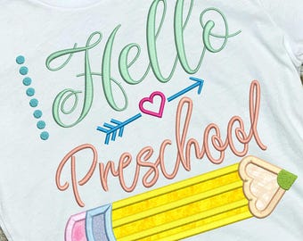 May include: White t-shirt with the words "Hello Preschool" in colorful script and a pencil graphic. The design includes a teal arrow, a pink heart, and teal dots. The shirt is displayed on a woven basket.