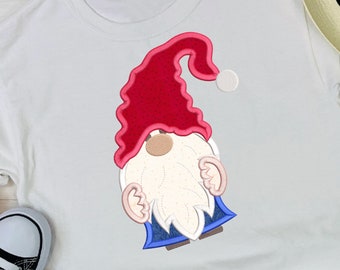 Könnte beinhalten: Weißes T-Shirt mit einem gestickten Gnom-Motiv. Der Gnom trägt einen roten Hut mit einem weißen Bommel, einen weißen Bart und eine blaue Robe.