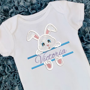 Peut inclure: Un body bébé blanc avec un lapin blanc pailleté qui regarde par-dessus une bannière bleue et blanche avec le nom "Victoria" en violet.