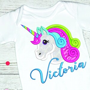 May include: White baby onesie with a colorful unicorn design. The unicorn has a silver body, a multi-colored mane, and a horn. The name "Victoria" is written in blue script below the unicorn.