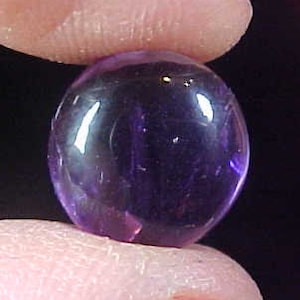 May include: A round, polished, deep purple gemstone, held between two fingers. The gem has a smooth, reflective surface, with light reflecting off its rounded form. The background is a dark, blurred gradient.