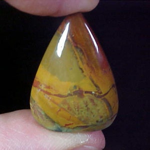 May include: A teardrop-shaped gemstone with a polished surface. The stone features a blend of colors, including yellow, brown, and red, with intricate patterns and veining. The gemstone is held between two fingers against a dark background.