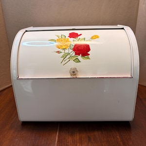 Peut inclure: Une boîte à pain en métal blanc avec une porte coulissante ornée d'un motif floral de roses rouges et jaunes avec des feuilles vertes. L'avant de la porte incurvée possède un bouton hexagonal transparent et facetté pour ouvrir le compartiment.