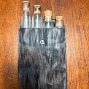May include: A black textured leather case containing four clear glass vials with cork stoppers. The case features a central silver-toned metal snap button closure. The vials are arranged in a row, with varying heights, and are partially visible above the top edge of the rectangular case.