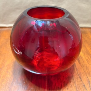 May include: A spherical red glass vase with a smooth, polished surface and a circular opening at the top. The translucent material allows light to pass through, creating deep crimson tones and reflections on the rounded body of the vessel.