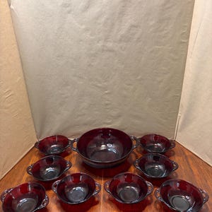 May include: A set of dark red glass tableware consisting of one large serving bowl and eight smaller matching bowls. Each bowl features two integrated side handles and a ribbed exterior texture, creating a uniform appearance across the collection.