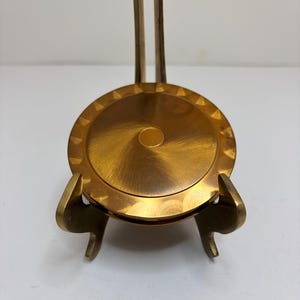 May include: A circular brass object with a brushed metallic finish and a central circular indentation, featuring a decorative scalloped edge. The object is mounted on a brass stand with two curved supports extending from the base to hold it upright.