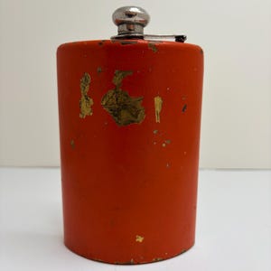 May include: A cylindrical orange metal flask featuring a silver-toned screw-top cap. The exterior surface displays significant wear, with multiple areas where the orange paint has chipped away to reveal the metallic base layer underneath.