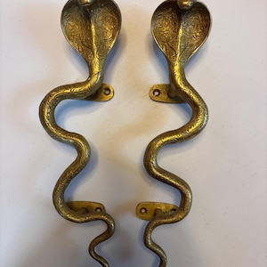 Könnte beinhalten: Zwei messingfarbene Türgriffe in Kobraform. Die Griffe sind mit geätzten Mustern versehen und haben eine goldene Oberfläche. Die Kobraköpfe sind ausgestellt, und die Körper krümmen sich in einer serpentinenartigen Form. Jeder Griff hat Montageplatten mit Schraubenlöchern.