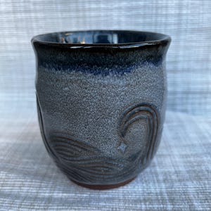 May include: A ceramic mug with a speckled gray and blue glaze, featuring a dark blue rim and a carved wave pattern on the exterior. The base of the mug is a reddish-brown clay color, and the overall shape is rounded with a wide opening.