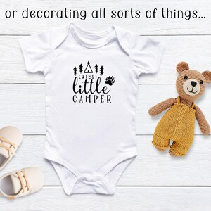 Puede incluir: Body blanco para beb&eacute; con el texto negro "Cutest Little Camper". El dise&ntilde;o incluye una tienda de campa&ntilde;a, &aacute;rboles y una huella de pata de oso. Un oso de peluche beige y zapatos de beb&eacute; tambi&eacute;n est&aacute;n en la imagen.