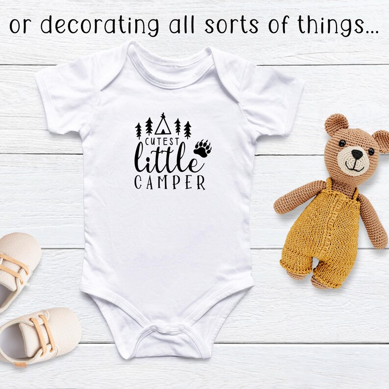 Puede incluir: Body blanco para beb&eacute; con el texto negro "Cutest Little Camper". El dise&ntilde;o incluye una tienda de campa&ntilde;a, &aacute;rboles y una huella de pata de oso. Un oso de peluche beige y zapatos de beb&eacute; tambi&eacute;n est&aacute;n en la imagen.