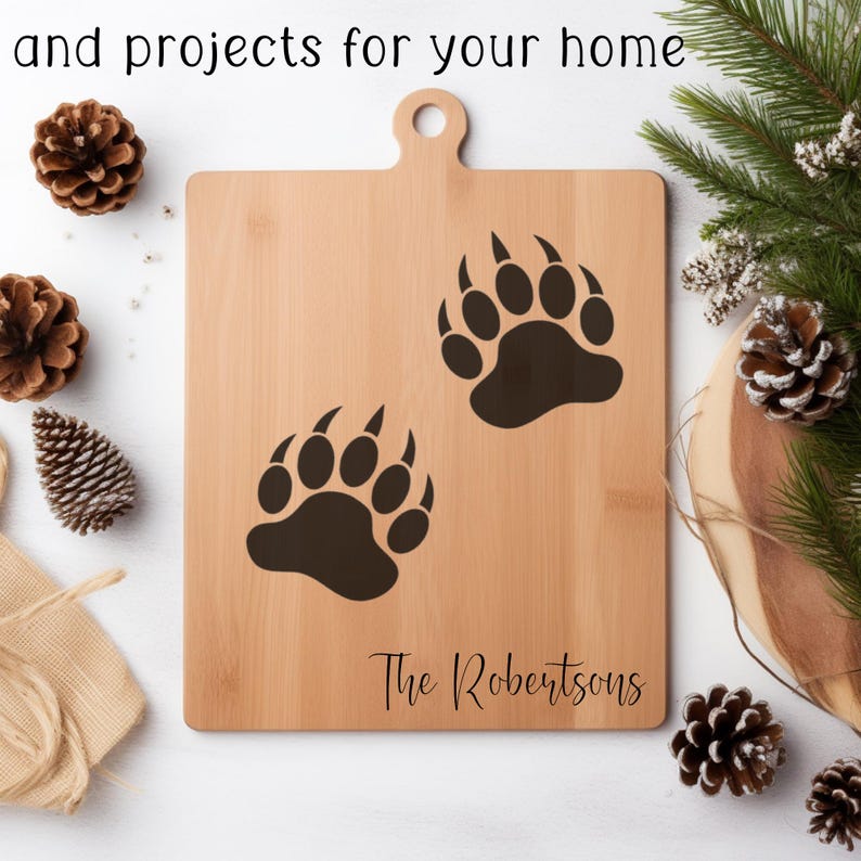 Puede incluir: Una tabla de cortar de madera marr&oacute;n claro con un asa y dos huellas de patas de oso negras. El texto "The Robertsons" est&aacute; impreso en escritura negra en la parte inferior. El texto "and projects for your home" est&aacute; en la parte superior.