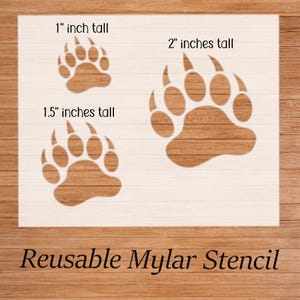 Puede incluir: Un est&eacute;ncil reutilizable de mylar blanco con tres huellas de oso de diferentes tama&ntilde;os: 2,5 cm, 3,8 cm y 5 cm de alto. Los est&eacute;nciles se muestran sobre un fondo de madera. El texto de la imagen dice "Reusable Mylar Stencil."