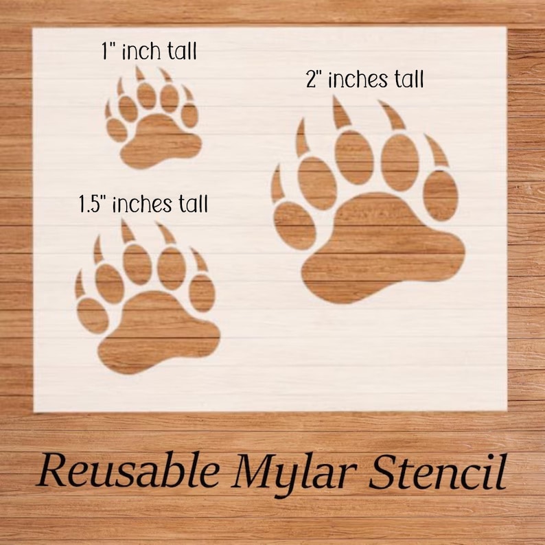 Puede incluir: Un est&eacute;ncil reutilizable de mylar blanco con tres huellas de oso de diferentes tama&ntilde;os: 2,5 cm, 3,8 cm y 5 cm de alto. Los est&eacute;nciles se muestran sobre un fondo de madera. El texto de la imagen dice "Reusable Mylar Stencil."