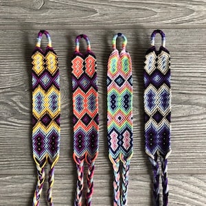 May include: Four colorful friendship bracelets with intricate geometric patterns in shades of blue, purple, yellow, orange, and white. The bracelets are made with braided string and have a loop at the top for easy wearing.