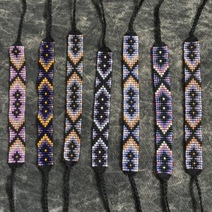 May include: Eight colorful beaded bracelets with geometric patterns in shades of purple, blue, black, white, and gold. The bracelets are tied with black cords.