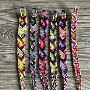 May include: Six colorful friendship bracelets with geometric patterns. The bracelets feature a variety of colors, including pink, green, purple, and black. Each bracelet has a loop at the top and braided ends.