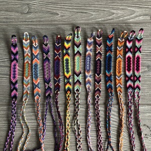 May include: A collection of 14 colorful friendship bracelets with various geometric patterns. The bracelets are made with colorful embroidery floss and are tied with a knot at the end.