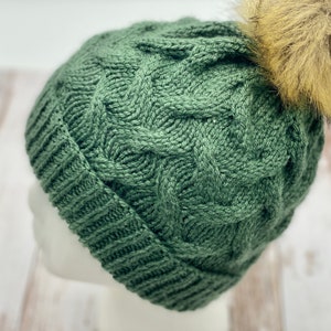 May include: A green knitted beanie with a cable knit pattern and a brown faux fur pom pom on top.