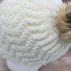 May include: White knitted beanie with a brown faux fur pom pom on top.
