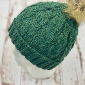May include: A green knitted beanie with a cable knit pattern and a fluffy brown pom pom on top.