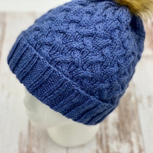 May include: A blue knitted beanie with a cable knit pattern and a fluffy pom pom on top.