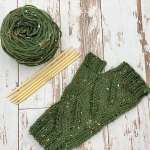 May include: A green and white knit fingerless glove with a cable knit pattern. The glove is lying on a white wooden surface with a ball of green yarn and knitting needles.