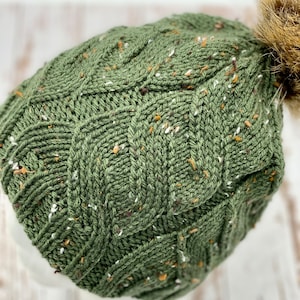 May include: A green knitted beanie with a cable knit pattern and a brown pom pom on top. The beanie has a speckled texture with white and brown flecks throughout.