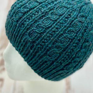 Op de afbeelding: Een teal en blauwe gebreide beanie-muts met een textuurpatroon. De muts is gemaakt van een zacht, warm materiaal en is ontworpen om nauwsluitend op het hoofd te passen. De beanie wordt getoond op een witte mannequinhoofd.