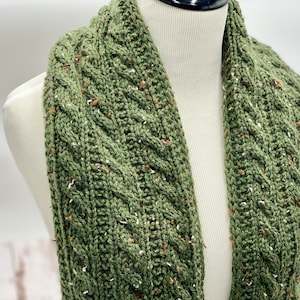 Può includere: Sciarpa verde lavorata a maglia con motivo a trecce. La sciarpa presenta macchie marroni e bianche nel filato. La sciarpa è esposta su un manichino. Un accessorio invernale.