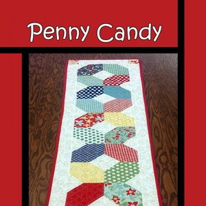 May include: A colorful table runner with a geometric pattern of hexagons in red, yellow, blue, green, and white. The runner is made of fabric with various patterns, including polka dots, floral prints, and plaid. The text "Penny Candy" is at the top of the image, and the text "Tricia Lynn Maloney Orphan Quilt Designs For Villa Rosa Designs" is at the bottom.