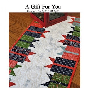 May include: A quilted table runner with a white center and colorful edges. The runner is 15.5 inches by 31.5 inches and features red, green, and navy blue patterned fabric. The text "A Gift For You" and "Creek Side Stitches" are also visible.