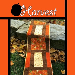 May include: A brown, orange, and cream quilted table runner with a harvest theme. The runner features a repeating pattern of pumpkins and leaves. The text "Harvest" is at the top of the image. The text "Tricia Lynn Maloney Orphan Quilt Designs For Villa Rosa Designs" is at the bottom of the image.