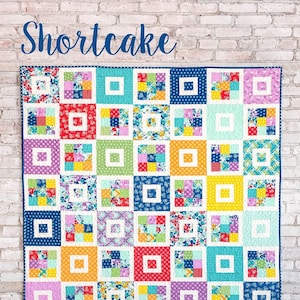 May include: A colorful patchwork quilt with the word "Shortcake" in script. The quilt features squares of various colors and patterns, including floral and polka dots. The text "A jelly roll quilt in 3 sizes" is also visible.
