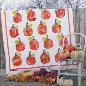 May include: A white quilt featuring a repeating pattern of orange and red patchwork pumpkins with green leaves, bordered by orange stripes. Several fabric pumpkins in various colors and textures are arranged on and around a white wooden chair.
