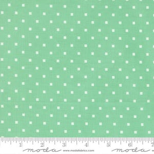 May include: A light green fabric featuring a repeating pattern of small white squares arranged in a grid. A white ruler is visible at the bottom edge, showing measurements in inches, along with the text moda www.modafabrics.com.