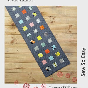 May include: A gray table runner with a patchwork design featuring small, colorful squares. The runner is quilted and has the text "RUNNER UP table runner" at the top. The design includes squares in orange, yellow, pink, and blue.